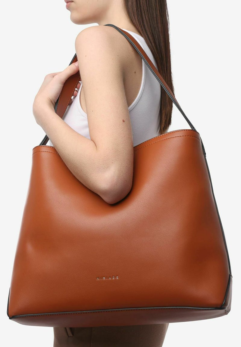 Borsa tote in pelle marrone con finitura liscia, grande corpo e rifiniture nere, tenuta da una persona. Logo in rilievo sottile sul davanti.