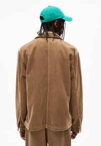 Brown corduroy jacket featuring a back seam, long sleeves, and a relaxed fit, paired with a green cap. Textured surface detail visible.