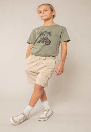 Kind staat met handen in zakken, draagt een groen T-shirt met motorfiets en palmboom print, beige korte broek, witte sokken en sneakers.