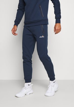 Mann trägt marineblaue Ellesse-Sweatpants und passendes Sweatshirt mit weißen Nike-Sneakern und Socken vor hellem, einfarbigem Hintergrund.