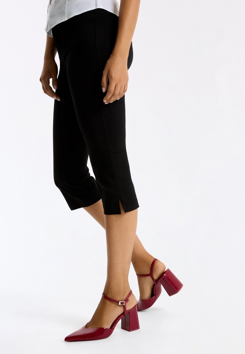 Femme portant un pantalon noir court avec des fentes latérales et des chaussures rouges à talon bloc, bout pointu et bride à la cheville sur fond blanc uni.