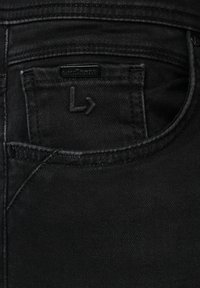 Czarne dżinsy z denimu z tylną kieszenią po prawej stronie, na której znajduje się subtelna naszywka z logo oraz dekoracyjne przeszycia wzdłuż pasa i krawędzi kieszeni.