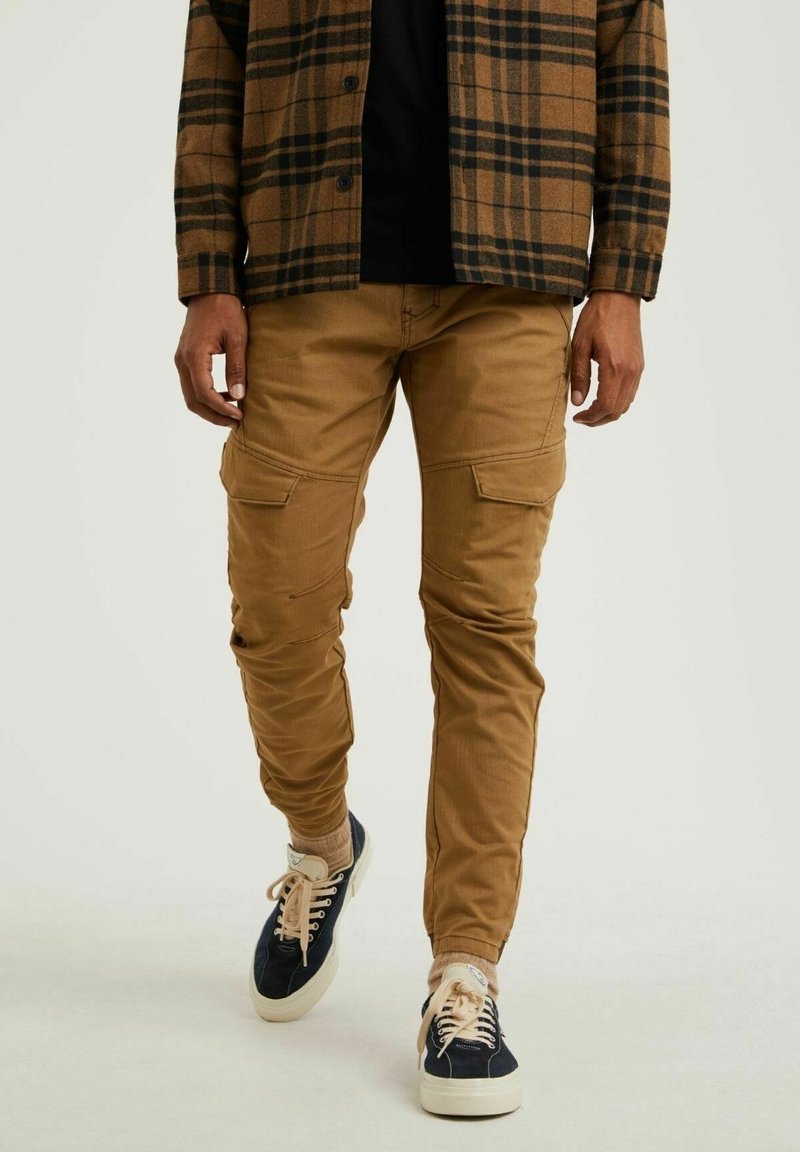 Homme portant un pantalon cargo marron, une chemise à carreaux noire et marron, des chaussettes beiges et des baskets noires marchant sur un fond blanc uni.