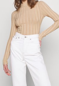 Beige lange mouw geribde top met een hoge kraag, gecombineerd met hooggepaste witte jeans met voorzakken en een metalen knoopsluiting.