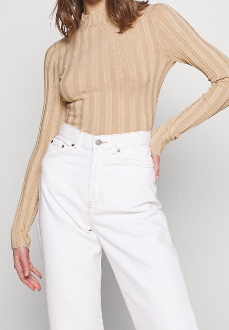 Beige lange mouw geribde top met een hoge kraag, gecombineerd met hooggepaste witte jeans met voorzakken en een metalen knoopsluiting.