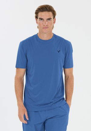 Ung mand med lys hud og tilbagelagt hår iført en ensfarvet blå t-shirt og matchende blå bukser, stående mod en hvid baggrund.