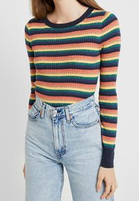 Ribbineuloksinen, pitkähihainen neule monivärisillä raidoilla (punainen, keltainen, vihreä, sininen) ja tummansininen pääntie, yhdistetty korkeavyötäröisiin, vaaleanpesun farkkuihin.