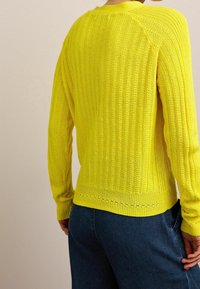 Maglione giallo lavorato a maglia con texture a costine, maniche lunghe e scollatura rotonda, con piccoli dettagli decorativi a occhio al bordo.
