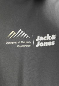 Grå stof med en glat tekstur, der har hvid og cremefarvet tekst samt et bjergmotiv. Teksten lyder "Designet på The Islet, København" og "Jack & Jones."