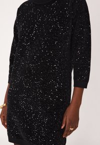 Abito nero in paillettes con una morbida texture, scollatura rotonda e maniche a tre quarti. Le paillettes creano un effetto scintillante in tutto l'abito.