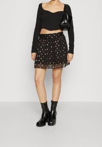 Svart långärmad crop top i kombination med en svart kjol som har ett genomskinligt överdrag och detaljer i guldbladsmönster. Svarta ankelstövlar fulländar looken.