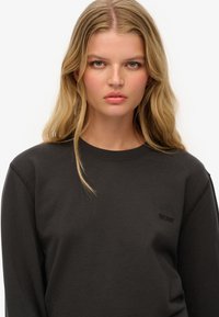 Joven mujer con cabello largo y rubio, que lleva una sudadera negra con un pequeño logo bordado de "Superdry" en el pecho, fondo neutro.