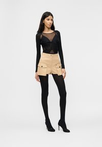 Γυναίκα που φοράει μαύρο μακρυμάνικο τοπ από διχτυωτό ύφασμα, μπεζ σορτς με ψηλή μέση και μεγάλες τσέπες, μαύρες καλσόν και μαύρα παπούτσια με ψηλά τακούνια σε λευκό φόντο.