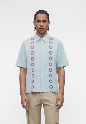 Άνδρας με πλεξουδάκια, που φοράει ανοιχτομπλε κοντομάνικο πουκάμισο με κάθετα μοβ κυκλικά μοτίβα και μπεζ παντελόνι σε λευκό φόντο.