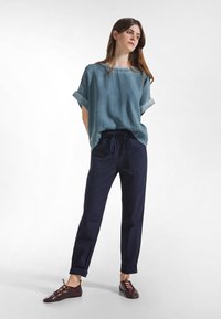 Jasnoniebieska lniana bluzka z podwiniętymi rękawami, granatowe spodnie z ściągaczem oraz ciemnobrązowe sznurowane buty, stojące na białym tle.