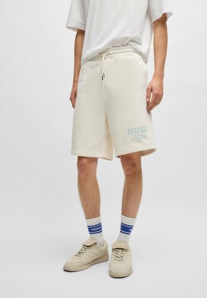 Personne portant un short beige clair jusqu'au genou avec le logo "HUGO", un T-shirt blanc, des chaussettes blanches à rayures bleues et des baskets beiges.