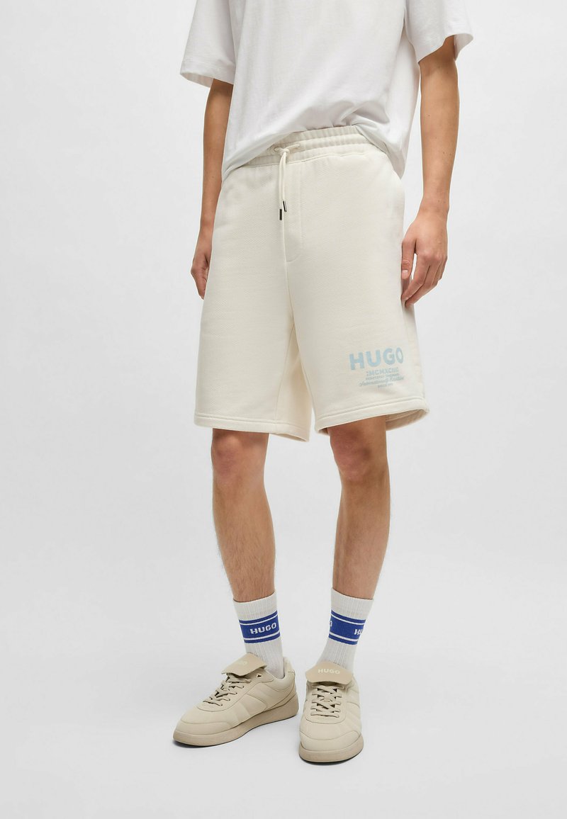 Personne portant un short beige clair jusqu'au genou avec le logo "HUGO", un T-shirt blanc, des chaussettes blanches à rayures bleues et des baskets beiges.