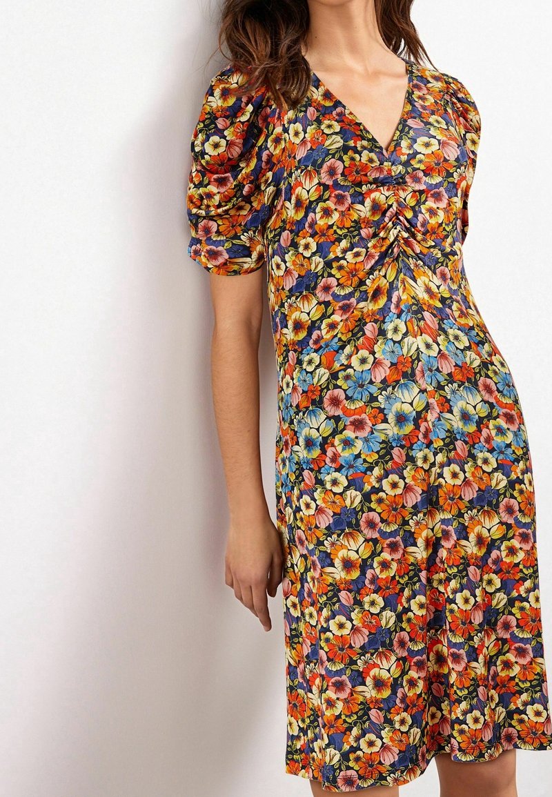 Robe fleurie avec un fond bleu marine et des fleurs multicolores en orange, jaune et bleu. Encolure en V et manches bouffantes. Longueur au-dessus du genou.