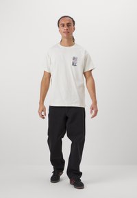 Cremfarbene Baumwoll-T-Shirt mit einem kleinen schwarzen Grafik auf der linken Brust. Getragen mit locker sitzenden schwarzen Jeans und schwarzen Schuhen mit roten Akzenten.