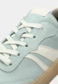 Ανδρικό αθλητικό παπούτσι από γαλάζιο σουέντ με λευκά κορδόνια και λευκή πλευρική ρίγα σε σχήμα κύματος, απεικονίζεται σε κοντινή λεπτομέρεια.