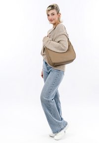 Sac à main en cuir tan avec une texture lisse et un design incurvé, comportant une fermeture éclair latérale et un détail de logo. Le modèle porte un pull beige et un jean bleu.