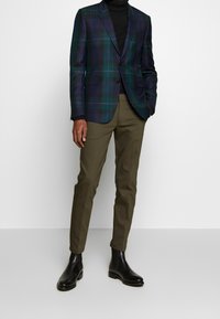 Blazer a quadri in lana verde scuro e blu navy, monopetto con rever a punta. Pantaloni slim-fit oliva e stivaletti in pelle nera.