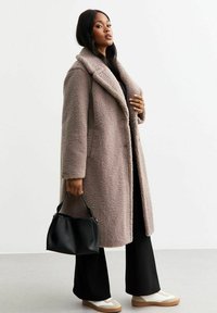 Cappotto in pelliccia sintetica color marrone chiaro, con un ampio collo e tasche laterali. Abbinato a pantaloni neri a gamba larga e sneakers color crema. Tiene una borsa nera.