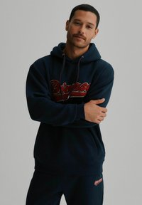 Granatowy hoodie z dużym haftowanym logo w kolorze czerwonym i białym. Posiada długie rękawy, kieszeń z przodu oraz sznurek do regulacji kaptura.