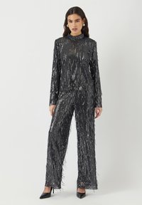 Sort paljettet outfit med en fransed langærmet bluse og brede bukser, med en tekstureret overflade og høj hals.