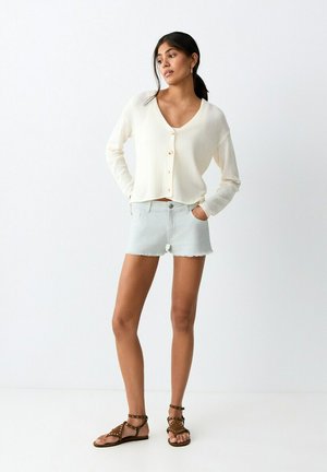 Jeune femme aux cheveux foncés attachés en queue de cheval, portant un cardigan crème boutonné, un short en jean clair et des sandales brunes cloutées, debout devant un fond blanc.