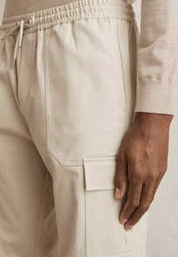 Pantalons cargo beiges avec une taille élastique et un cordon de serrage, comportant des poches latérales et une poche à patch sur la cuisse. Texture de tissu lisse.