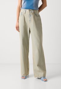 Jean large bleu clair en denim, avec une taille haute, cinq poches et un léger dégradé pour une coupe décontractée. Associé à des sandales bleues.