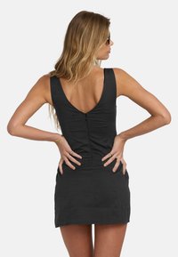 Woman with long blonde hair wearing sunglasses and a sleeveless black dress with a low back and zipper, hands resting on hips.
