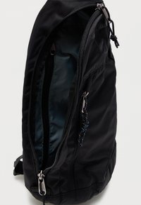 Μαύρο σακίδιο πλάτης με μερικώς ανοιχτό κύριο φερμουάρ που δείχνει τη σκοτεινή εσωτερική επένδυση και μπροστινή τσέπη με φερμουάρ, με μαύρα και μπλε κορδόνια έλξης.