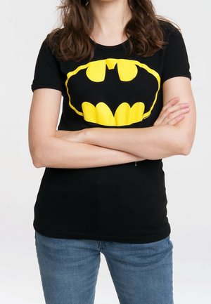 Woman with folded arms wearing black t-shirt featuring large yellow Batman logo and blue jeans against plain background.