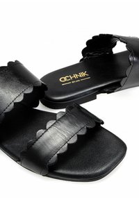 Czarne skórzane sandały z falistymi paskami, płaską podeszwą i gładką fakturą. Posiadają logo na wkładce w złotym kolorze.