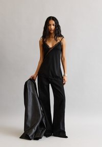 Hosszú, sötét hajú nő fekete csipkés pántos felsőt és bőszárú nadrágot visel, egy fekete bőrdzsekit tart a háttér előtt.