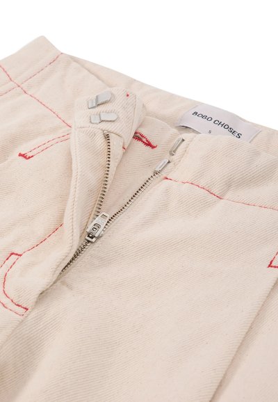 Pantalon beige avec coutures rouges, fermeture éclair avant partiellement ouverte, fermeture métallique à crochet et barre, et une étiquette de marque visible indiquant Bobo Choses taille small.