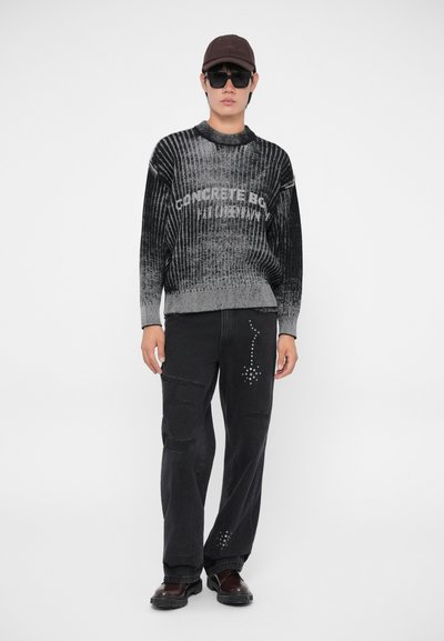 Un suéter de punto con degradado en negro y gris presenta una textura acanalada y texto impreso, combinado con jeans negros que tienen detalles de tachuelas y desgarros.