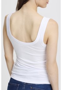 Sieviete redzama no aizmugures, valkājot baltu bezroku topu ar platām siksnām un zilas džinsas uz gaiša vienkārša fona.