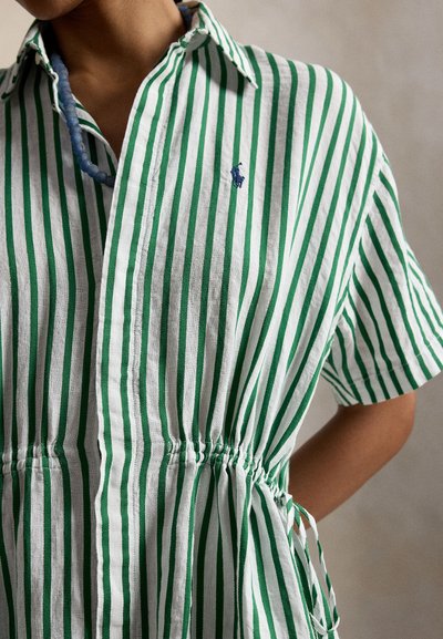 Persona che indossa una maglietta a maniche corte con righe verticali verdi e bianche, vita stretta, e una collana di perline blu.