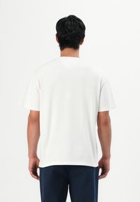 Man with dark hair wearing a plain white short-sleeve T-shirt and dark pants, facing away against a white background.