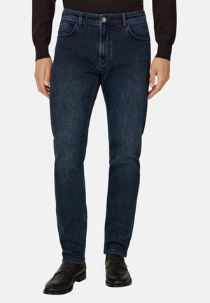 Mand iført mørkeblå slim-fit jeans, sort langærmet skjorte og sorte læder loafers, stående mod en ensfarvet baggrund.