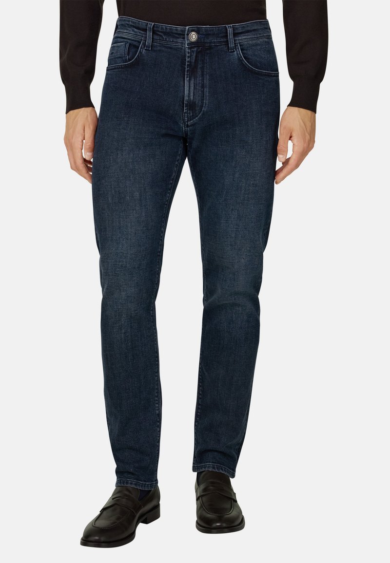 Homme portant un jean slim bleu foncé, une chemise noire à manches longues et des mocassins en cuir noirs, debout devant un fond uni.