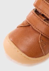 Καφέ δερμάτινο παπούτσι με δύο ρυθμιζόμενα λουριά, στρογγυλερή μύτη και λαστιχένια σόλα. Υφή στο δέρμα. Ραμμένες λεπτομέρειες κοντά στα λουριά.