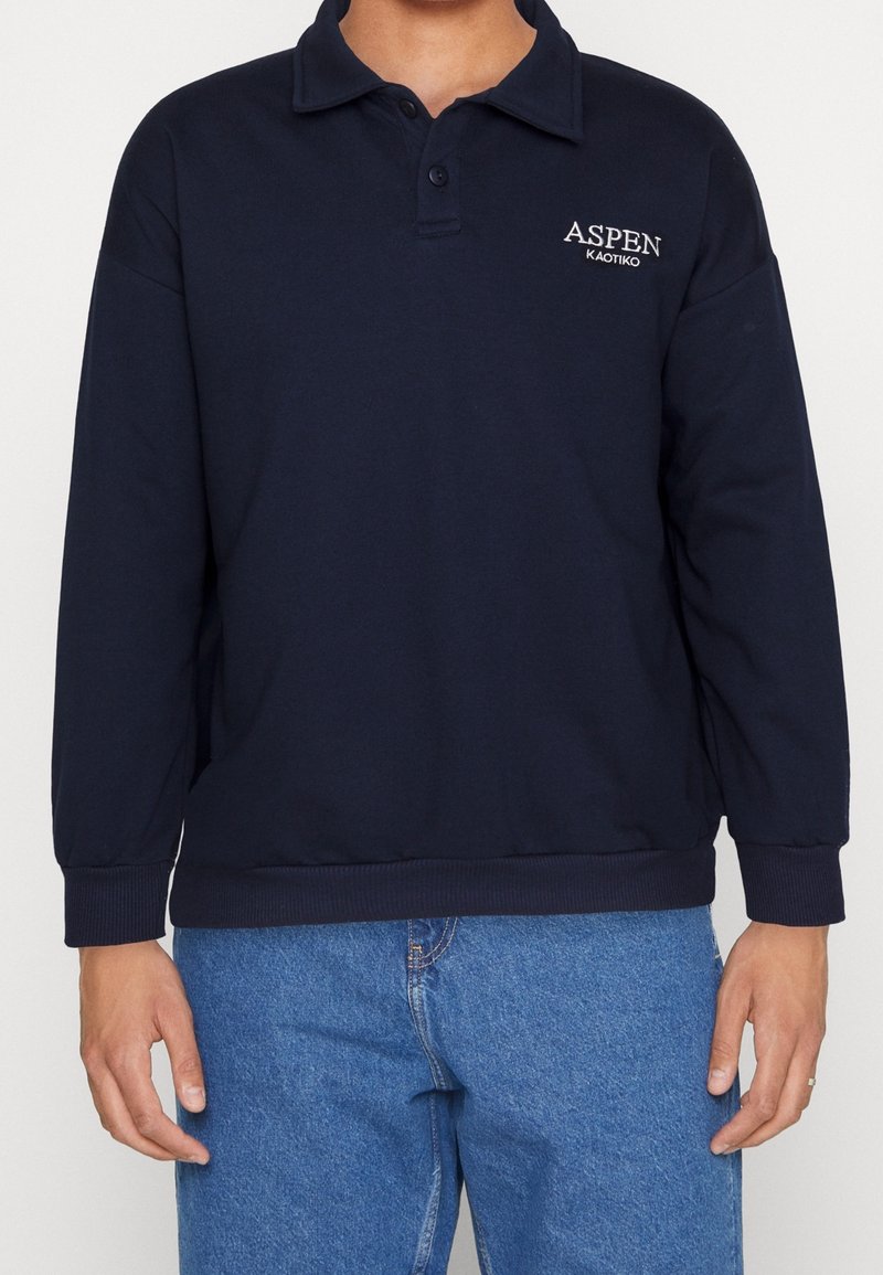 Man som bär en marinblå långärmad polo-tröja med "ASPEN KAOTIKO"-logotyp, i kombination med blå jeans, stående mot en enfärgad bakgrund.