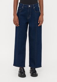 Jeans de perna larga em azul escuro, feitos de denim, com costuras em contraste, cinco bolsos e fecho em botão de metal. Bolsos frontais e traseiros planos.