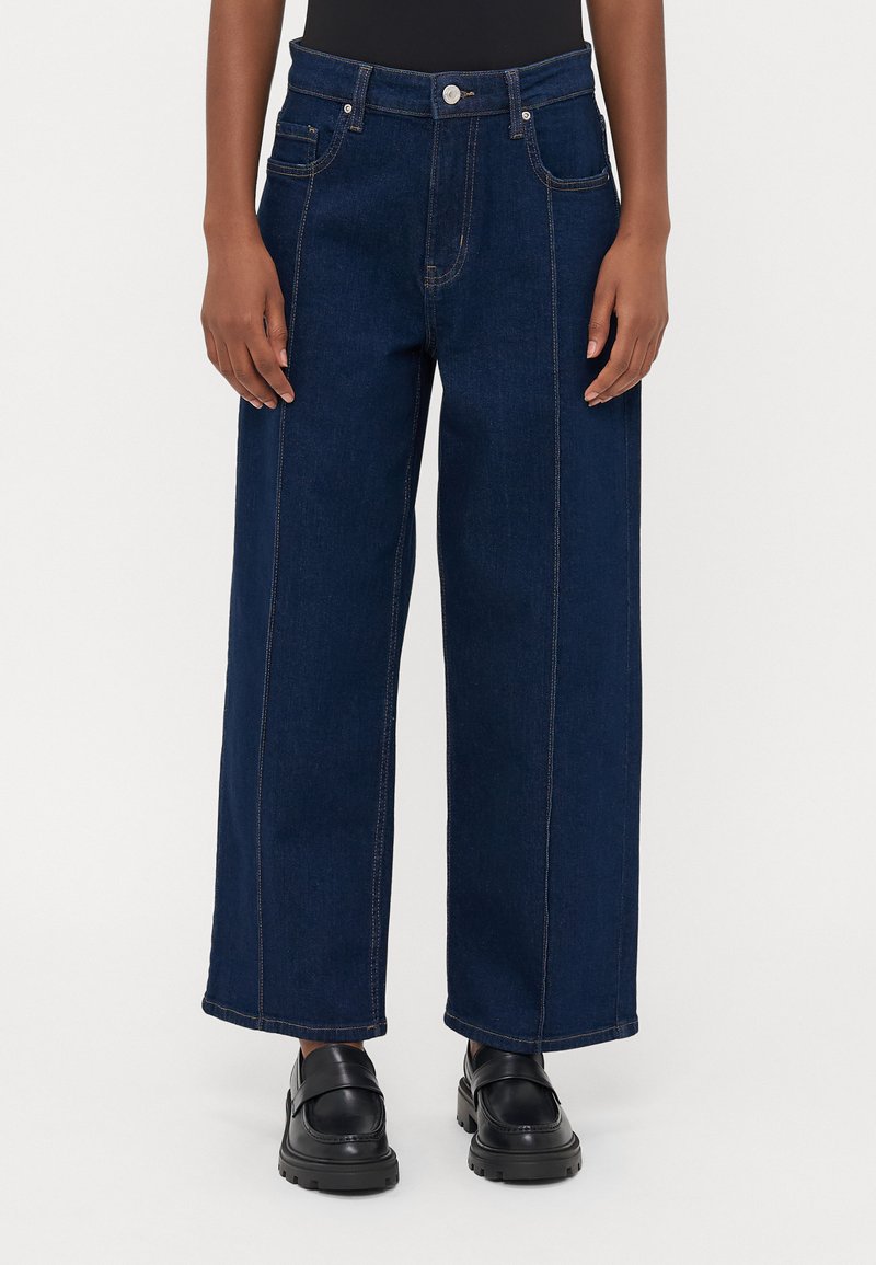 Jeans de perna larga em azul escuro, feitos de denim, com costuras em contraste, cinco bolsos e fecho em botão de metal. Bolsos frontais e traseiros planos.