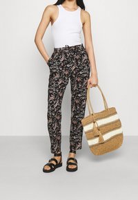 Black patterned trousers with floral design, paired with a white tank top and black sandals. Holding a woven bag with striped accents.