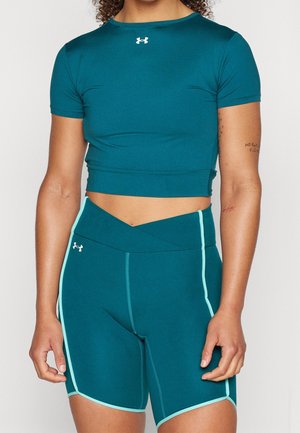 Kobieta ubrana w turkusowy krótki top Under Armour z krótkim rękawem i dopasowane sportowe szorty z wysokim stanem z jasnoniebieskimi wstawkami.